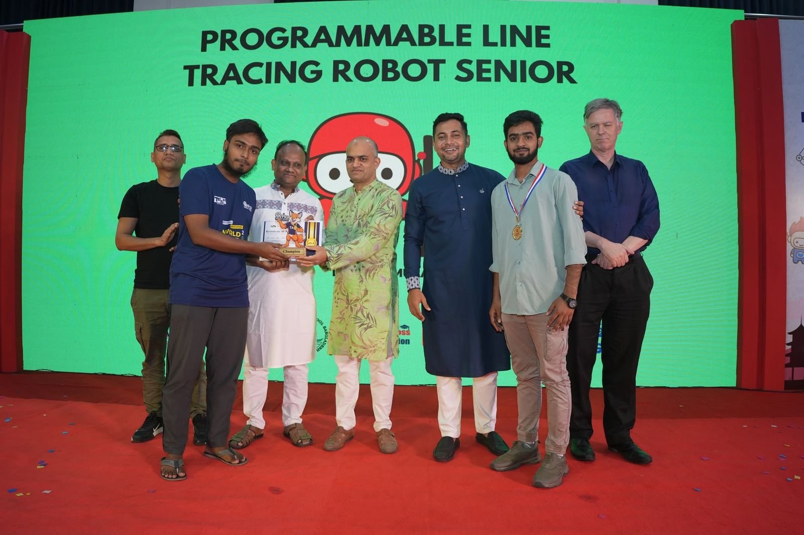 A Programmable Line Tracing Robot Senior winner receives a trophy on stage beside guests and a fellow medalist