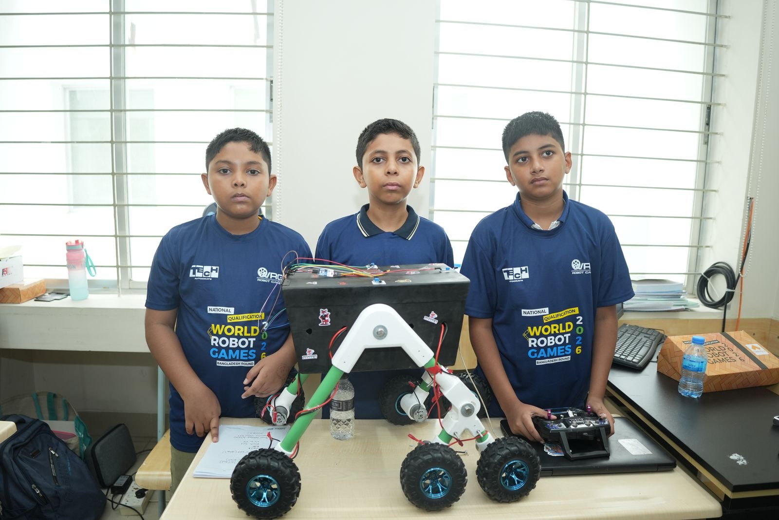Three junior participants stand behind their wheeled rover robot and its remote controller