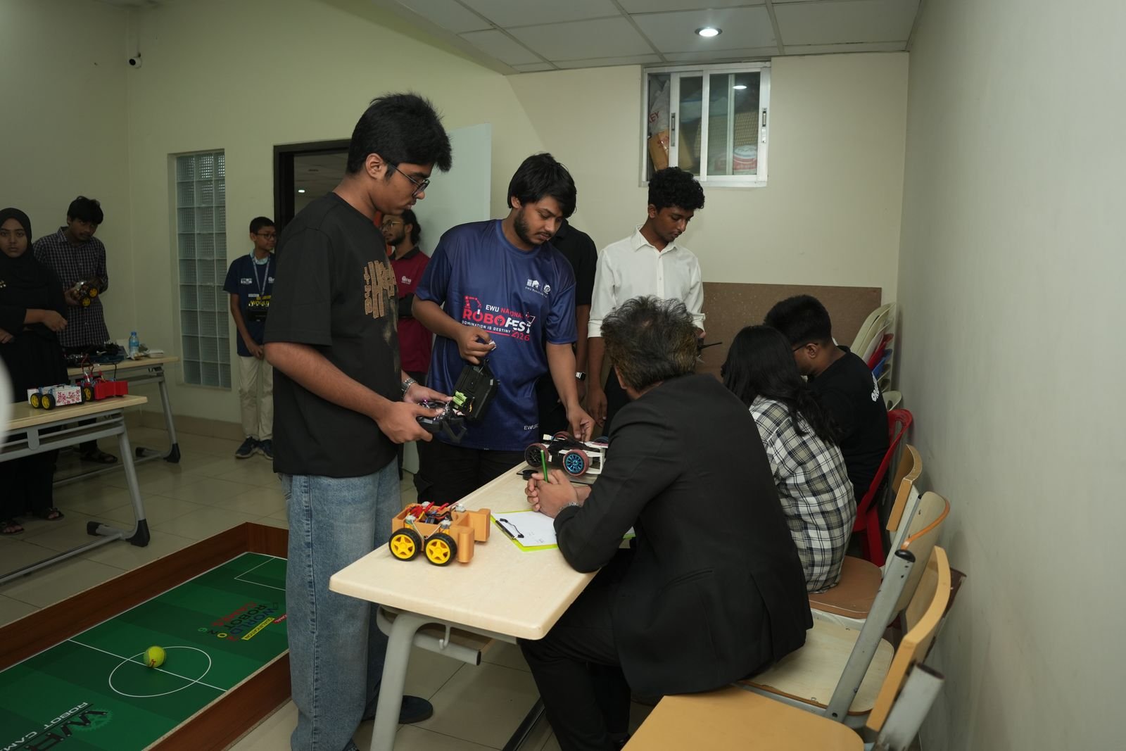 A participant drives a soccer robot by remote while a judge scores at the desk beside the field mat