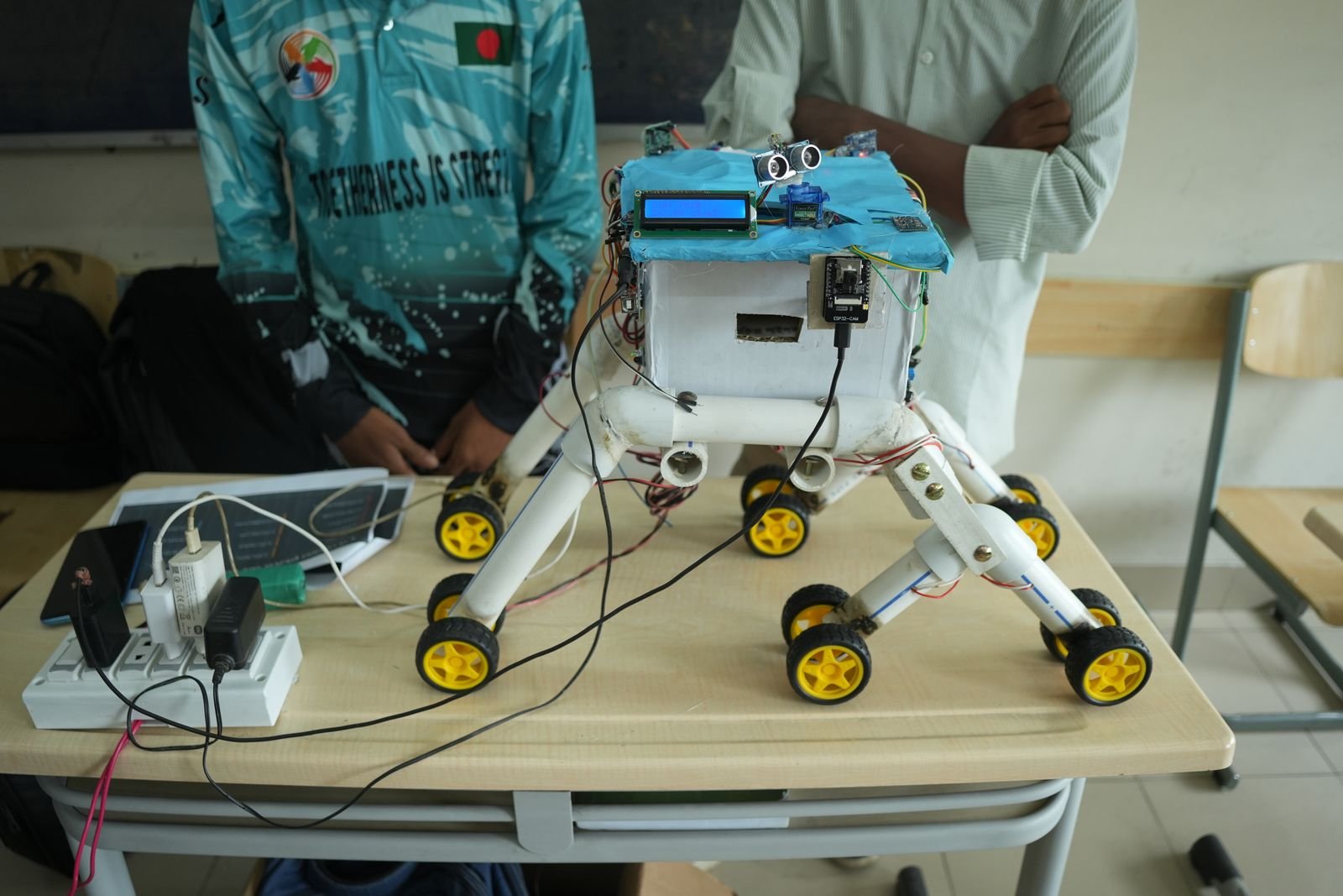 A multi-wheeled rover robot with an LCD screen and ultrasonic sensor on a classroom desk