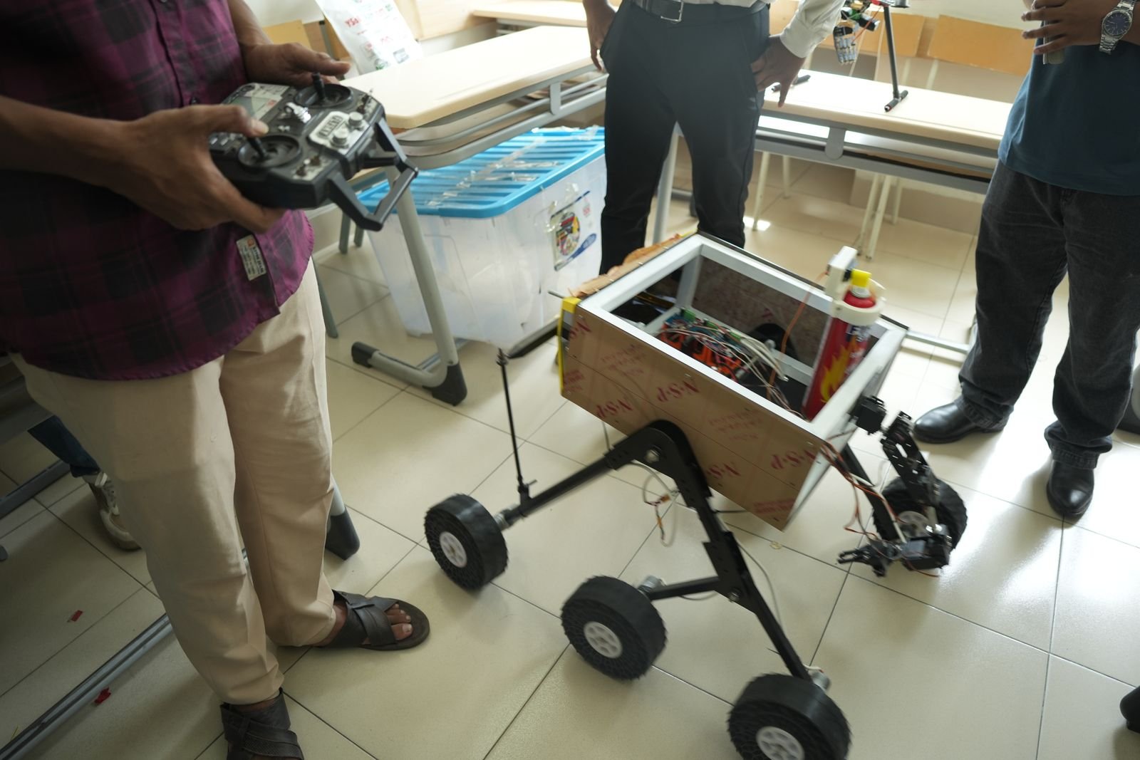 A participant holds a controller beside a wood-bodied rover fitted with a robotic arm