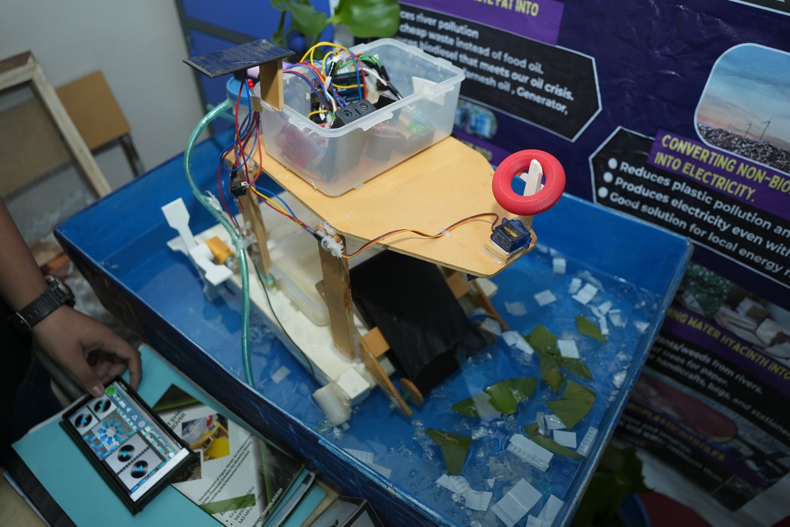 A water-tank prototype with a solar panel and sensors, displayed in front of its clean-energy project poster