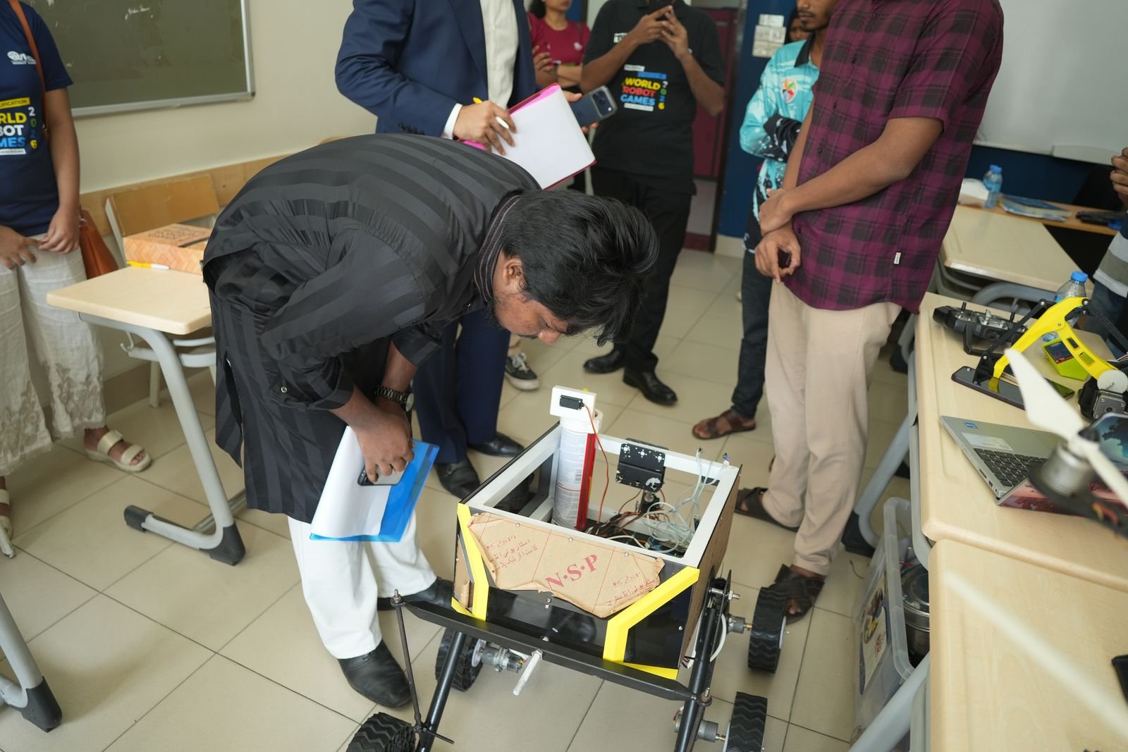 A judge bends down to inspect a tracked robot chassis on the classroom floor