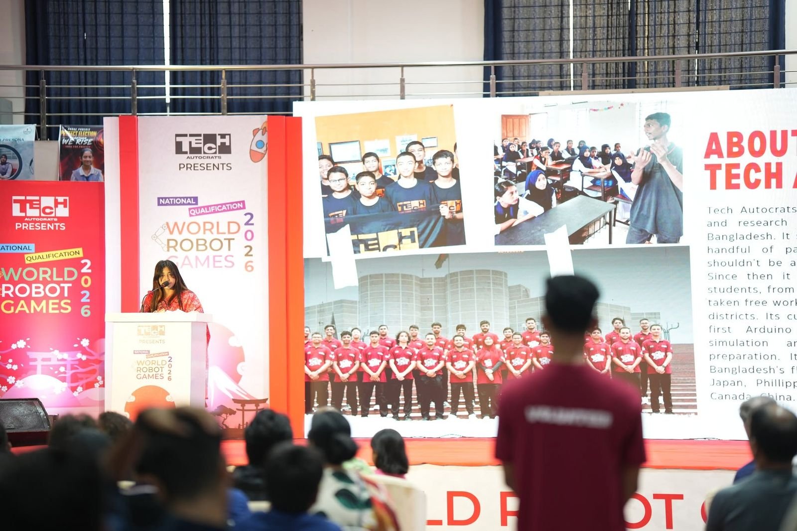 A host speaks from the podium beside an About Tech Autocrats display as the audience watches